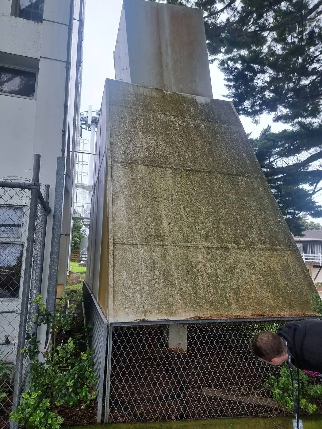 Moss-covered asbestos-cement ventilation hood on a steel frame beside a commercial building in Auckland, before SES removal